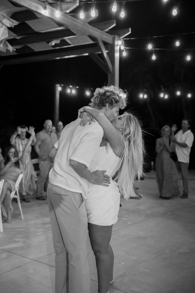 Couple embracing and kissing under string lights at night with guests clapping in background