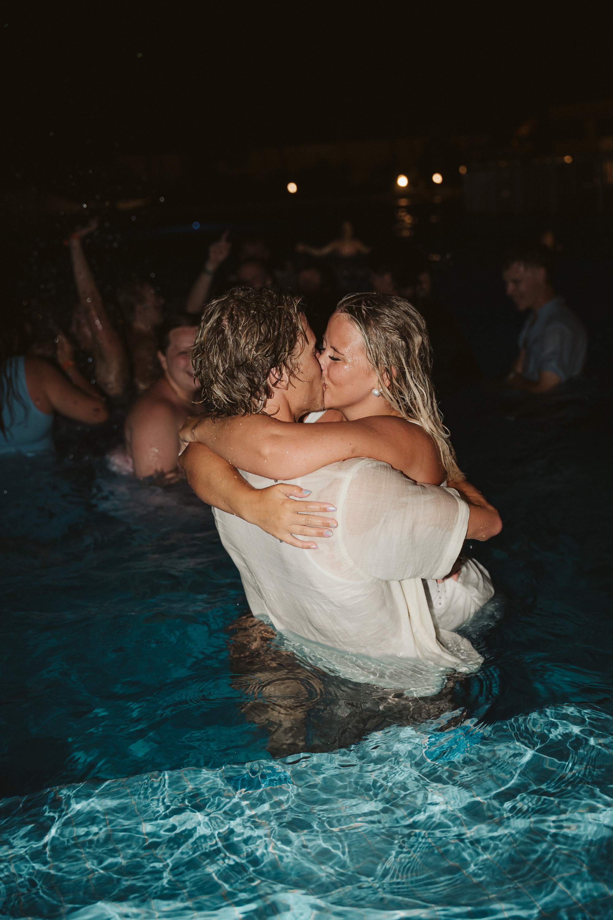 Newlyweds sharing a kiss in the pool surrounded by cheering wedding guests.